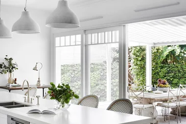 Bright modern kitchen with white countertops, pendant lights, and large windows overlooking a lush garden patio.