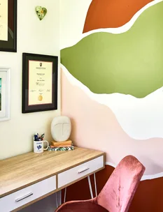 Cozy home office corner with abstract wall art, framed diploma, pink chair, and a desk with a mug and sculptural decor.