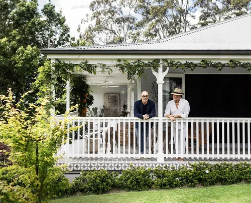 Two people leaning on a white verandah railing of a house surrounded by greenery.