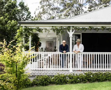 Two people leaning on a white verandah railing of a house surrounded by greenery.