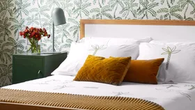 Cozy bedroom with green palm wallpaper, mustard pillows, and a green nightstand with flowers in a vase.