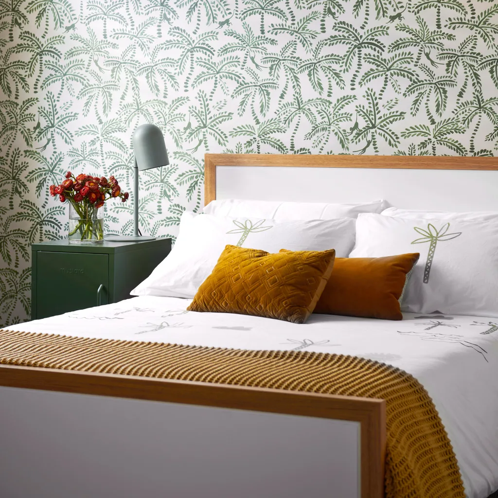 Cozy bedroom with green palm wallpaper, mustard pillows, and a green nightstand with flowers in a vase.
