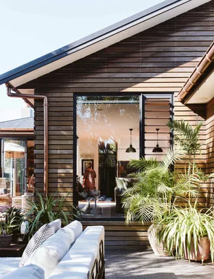 A modern wooden house exterior with large windows, outdoor seating, and lush potted plants under a clear blue sky.