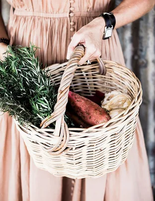 A person in a dress holds a wicker basket with rosemary, sweet potatoes, and garlic.