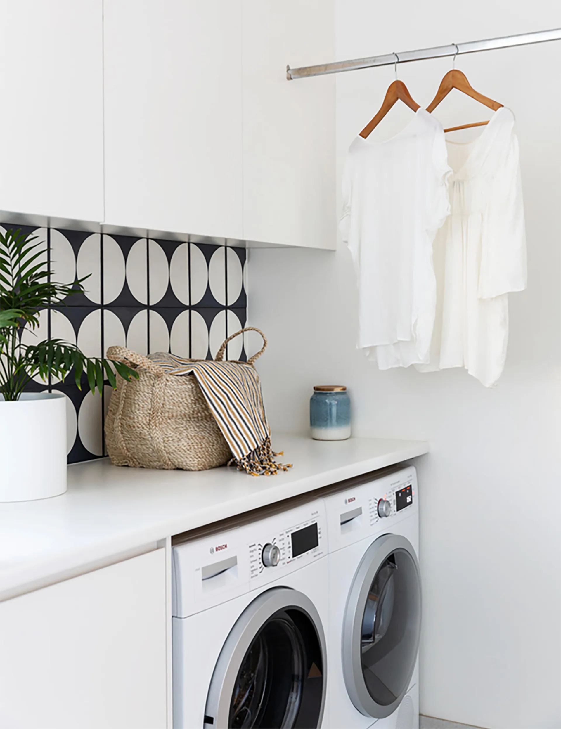 Laundry room with washer, dryer, patterned backsplash, plants, basket, and clothes hanging on a rod.