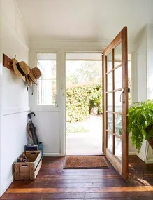 Front door open to a sunlit garden; hats on hooks, umbrella and shoes in a wooden crate on a polished wooden floor.