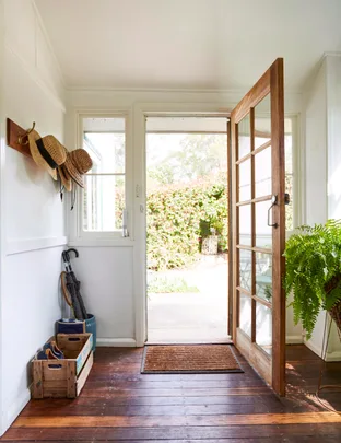 Front door open to a sunlit garden; hats on hooks, umbrella and shoes in a wooden crate on a polished wooden floor.