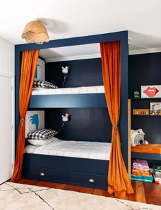Bunk beds with navy frame and orange curtains, checkered pillows, wall lamps, and colorful decor in a cozy room.