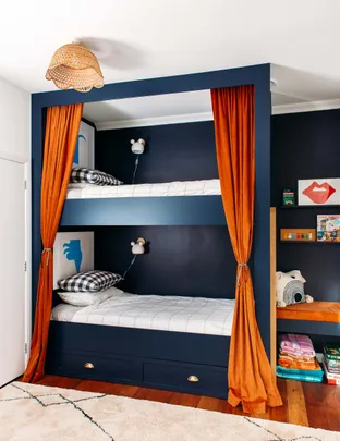 Bunk beds with navy frame and orange curtains, checkered pillows, wall lamps, and colorful decor in a cozy room.