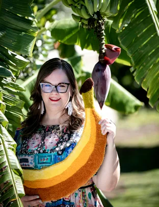 Person holding a large banana-shaped rug standing among banana trees.