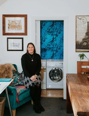 A woman sits on a teal chair in a cozy room with art on the walls, next to a washing machine alcove.