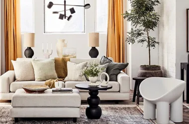 Modern living room with beige sofa, various pillows, a round table with flowers, a floor lamp, and a large potted plant.