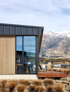 Modern wood-paneled house with large windows, people inside at a table, mountain view in the background.