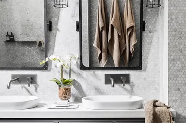 Modern bathroom with dual sinks, wall-mounted faucets, mirrors, hanging towels, and an orchid plant on a hexagonal tile wall.