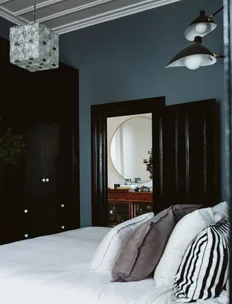 Cozy bedroom with a white bed, grey and striped pillows, dark walls, a modern light fixture, and a mirror in the background.