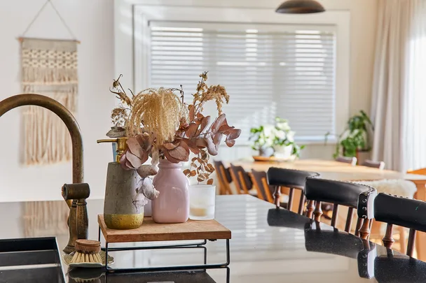 Dried flowers in vases on a kitchen counter, with a dining table and plants in the background.