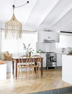 A bright kitchen with white walls, wooden chairs, a table, and a woven pendant light hanging from a high ceiling.