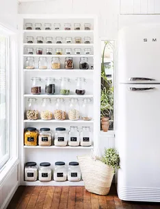 Clear glass jars with labeled ingredients on white shelves next to a vintage-style fridge.