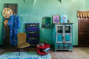 Colorful room with green walls, patterned textiles, a blue robe, wicker hats, vintage furniture, art, and a red table.