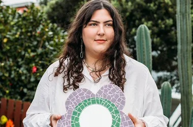 Person with long dark hair holding a flower-shaped, mosaic mirror, standing in a garden with cacti and greenery.