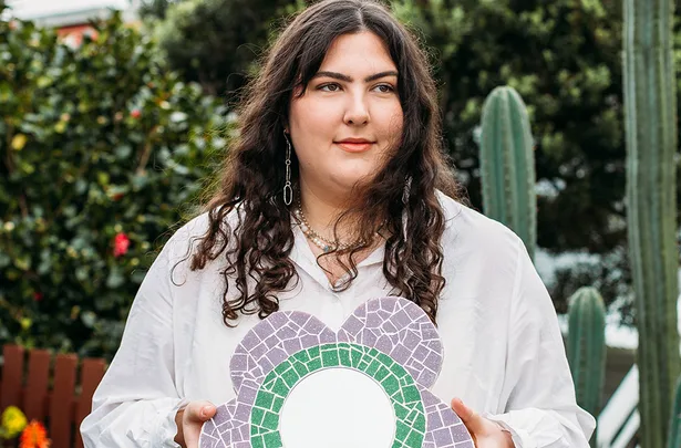 Person with long dark hair holding a flower-shaped, mosaic mirror, standing in a garden with cacti and greenery.