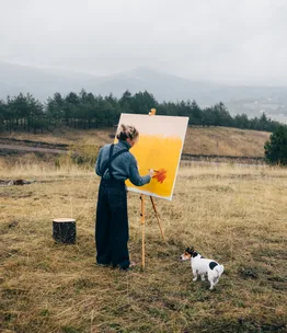Artist painting on canvas in a field, with a small dog beside them, surrounded by mountains and trees.