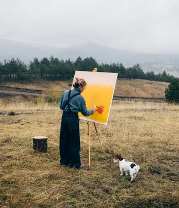 Artist painting on canvas in a field, with a small dog beside them, surrounded by mountains and trees.