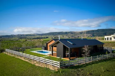 Modern house with black roof and pool in a green, fenced landscape under clear blue sky with hills in the background.