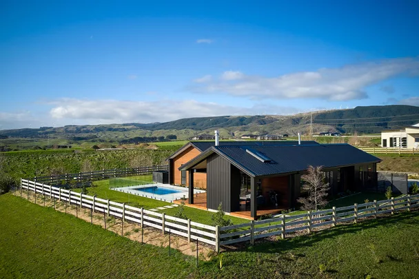 Modern house with black roof and pool in a green, fenced landscape under clear blue sky with hills in the background.
