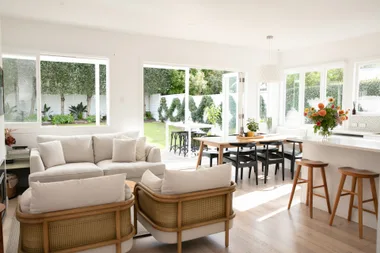 Bright living area with beige sofas, wood furniture, large windows, and open doors leading to a garden.