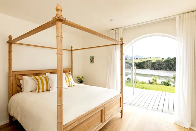 Four-poster bed with striped pillows in a bright room, open sliding doors to a river view and green landscape.