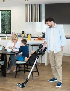 Man vacuuming kitchen floor while woman and child sit at table drawing.