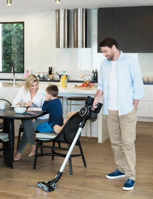 Man vacuuming kitchen floor while woman and child sit at table drawing.