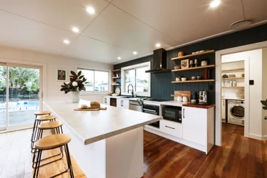 Modern kitchen with marble island, bar stools, open shelving, and view of laundry room. Large windows and sliding door.