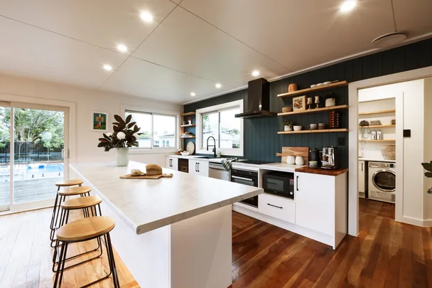 Modern kitchen with marble island, bar stools, open shelving, and view of laundry room. Large windows and sliding door.