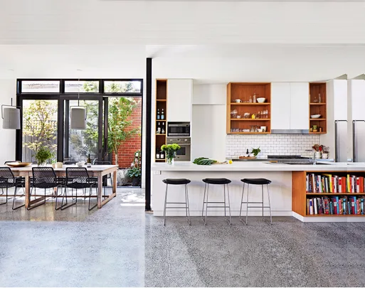 Modern kitchen with large windows, white cabinets, open shelves, dining table, and bar stools on a polished concrete floor.
