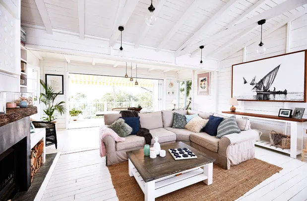 Bright living room with beige sectional, colorful cushions, wooden table, art on walls, and large windows opening to greenery.