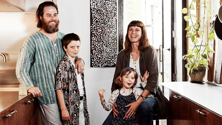 Family of four smiling in a modern kitchen with wooden cabinets and plants by the window.