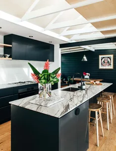 Modern kitchen with marble island, black cabinets, wooden stools, and tropical flowers in a vase.