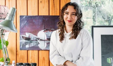 A person with curly hair in a white shirt smiles in an art studio with a painting of bread behind them.