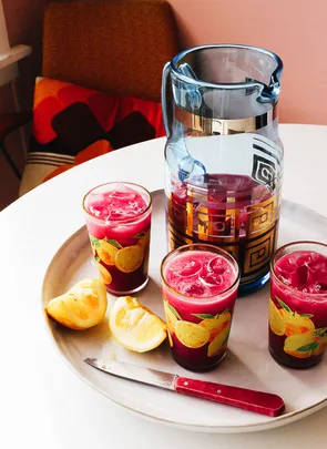 Pitcher and glasses of beet juice with ice, lemon slices, and knife on a table with a colorful cushion in the background.