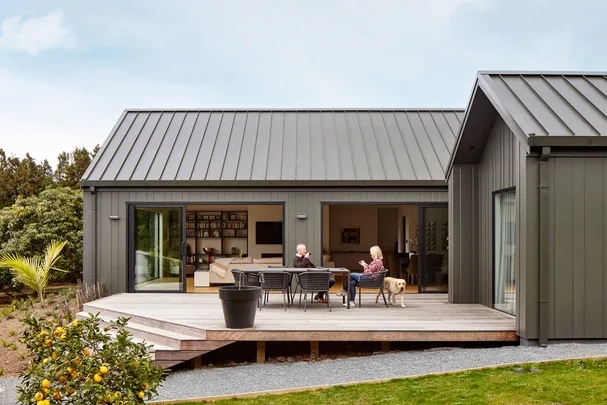 Outdoor patio with two people seated at a table, a dog nearby, and a modern house in the background.