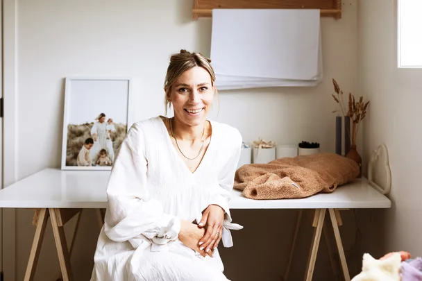 Woman in white dress smiling, sitting at a table with framed family photo and cozy decor items in a bright room.