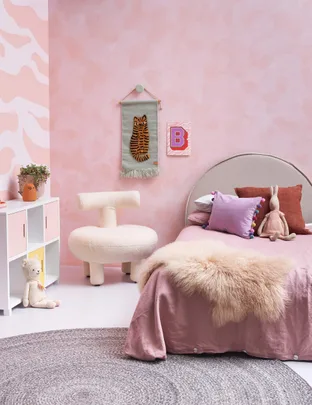 Cozy pink kids' room with colorful pillows, plush toys, and a wall tapestry of a tiger. Beige chair and round rug.