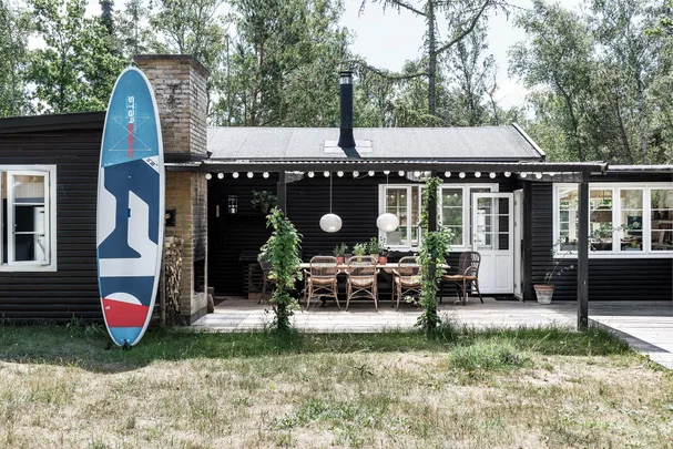 A cozy wooden cottage with a surfboard, outdoor dining area, and string lights, surrounded by lush greenery.