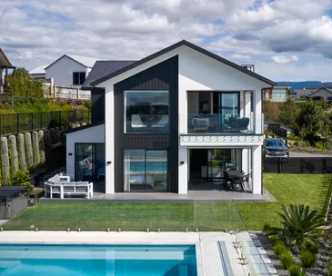 Modern two-story house with large windows, balcony, green lawn, and a pool in the foreground under cloudy skies.