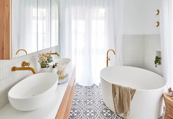 Elegant white bathroom with a freestanding tub, gold fixtures, patterned tile floor, and large windows with sheer curtains.
