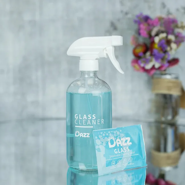 Glass cleaner spray bottle and dissolvable cleaning tablet packet on a reflective surface, flowers in the background.