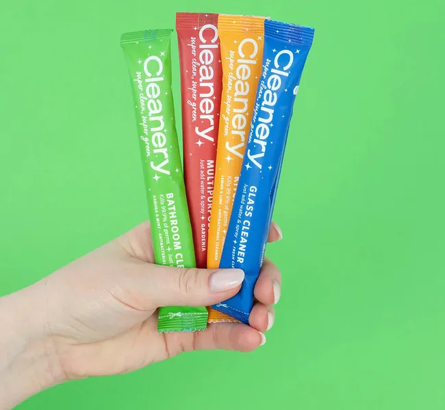 Hand holding four Cleanery cleaner packets against a green background.