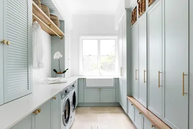 Bright and airy laundry room with light blue cabinets, washing machines, shelving, and a large window.